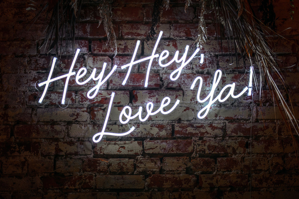 Et hvitt neonskilt med teksten «Hey Hey, Love Ya!» på en murvegg.